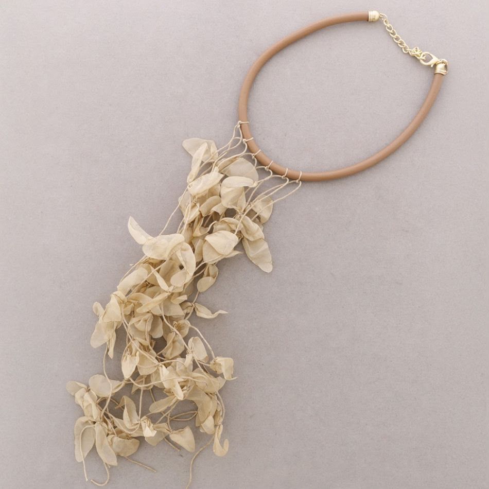 Collar beige con borla floral y cierre dorado sobre fondo gris claro.