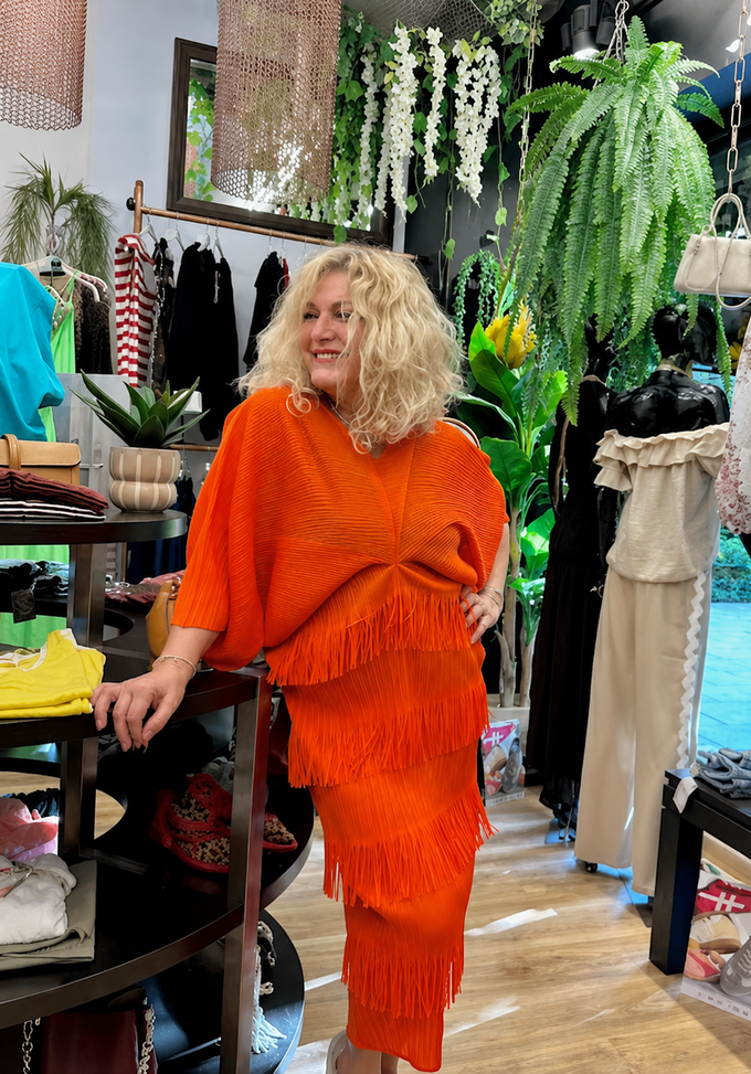 Mujer con un llamativo conjunto de flecos naranjas posando en una boutique rodeada de plantas colgantes y percheros.