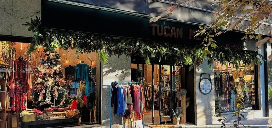 Escaparate de una tienda de ropa con prendas coloridas colgadas en el exterior bajo un toldo cubierto de enredaderas.