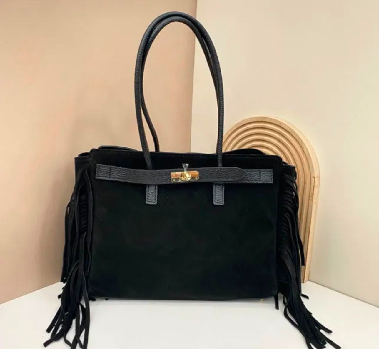 Bolso negro con borlas y cierre dorado sobre una superficie blanca sobre un fondo beige.