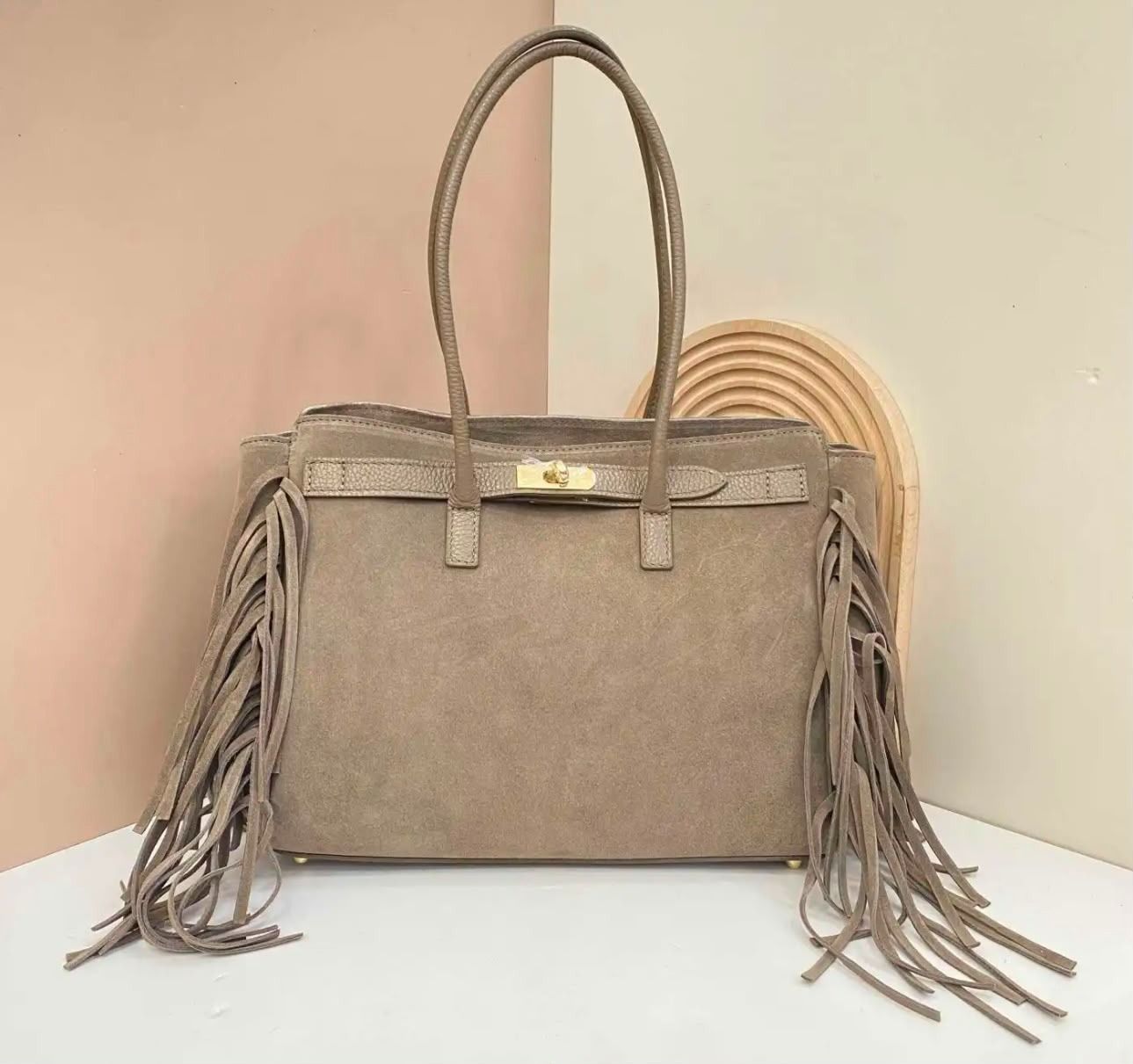 Bolso color topo con flecos largos en los laterales, apoyado verticalmente contra una pared lisa.