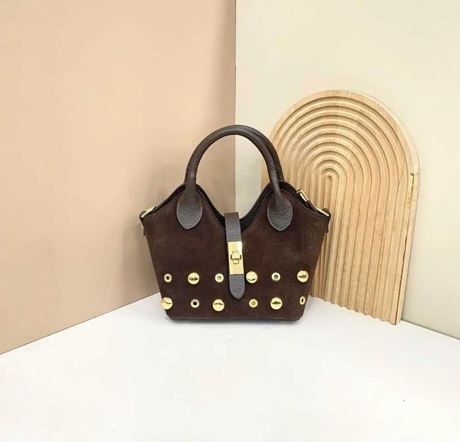 Bolso de mano marrón oscuro con tachuelas doradas y asa negra sobre una superficie blanca contra una pared beige.
