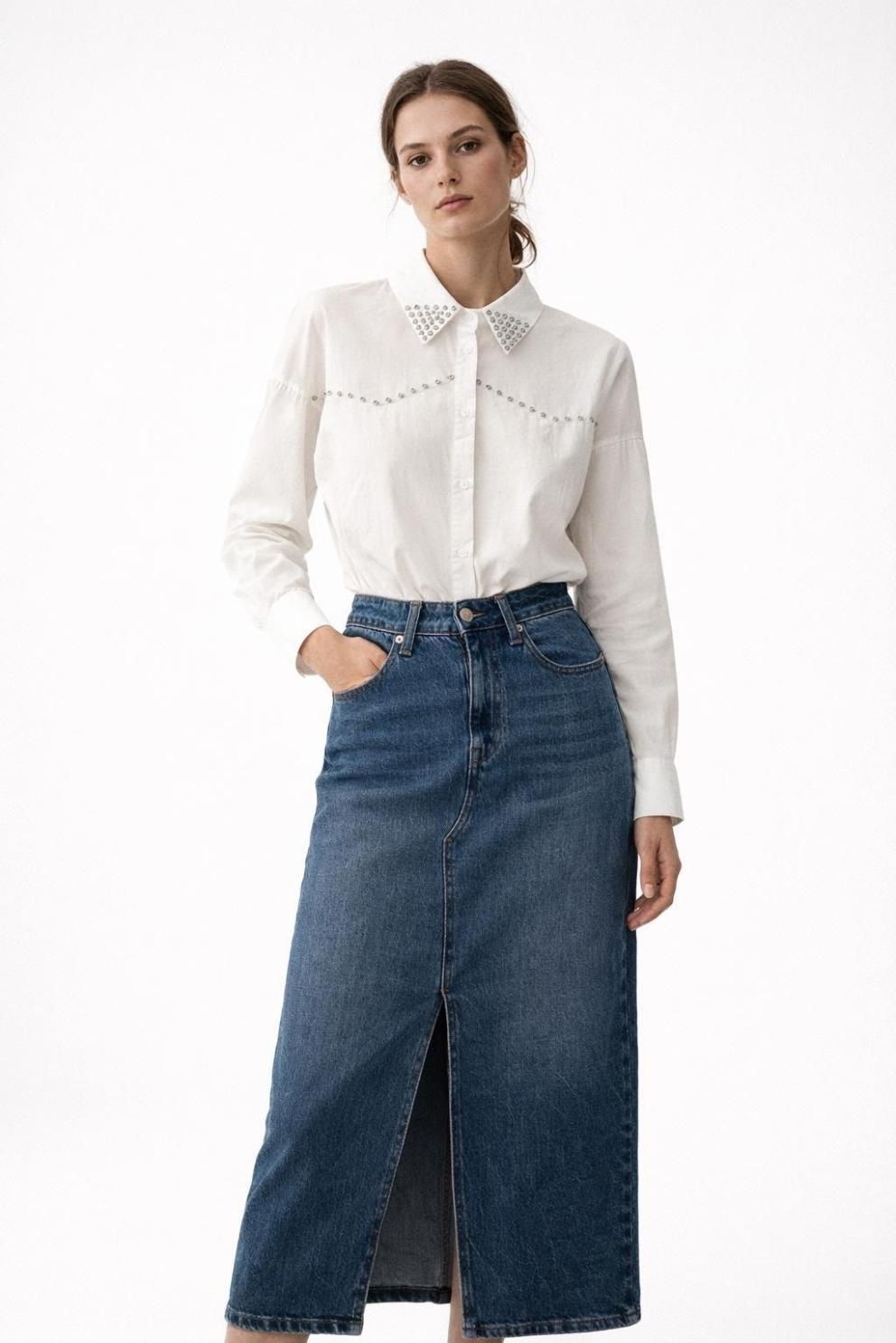 Una modelo luce una blusa blanca y una falda larga de mezclilla azul sobre un fondo liso.