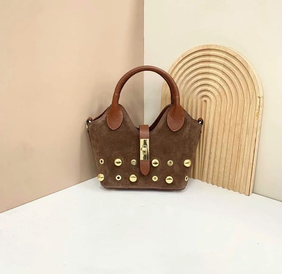 Bolso marrón con asas color canela y tachuelas doradas sobre una mesa blanca junto a un arco decorativo tejido.