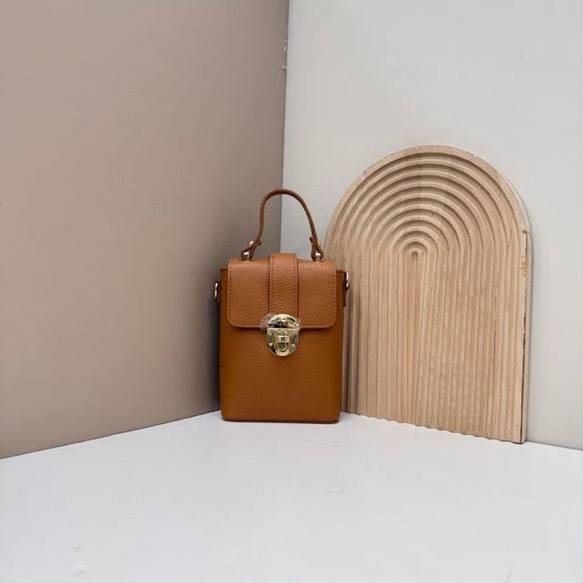 Bolso de mano de piel color camel con cierre dorado sobre una superficie blanca junto a una pieza decorativa de arco tejido.