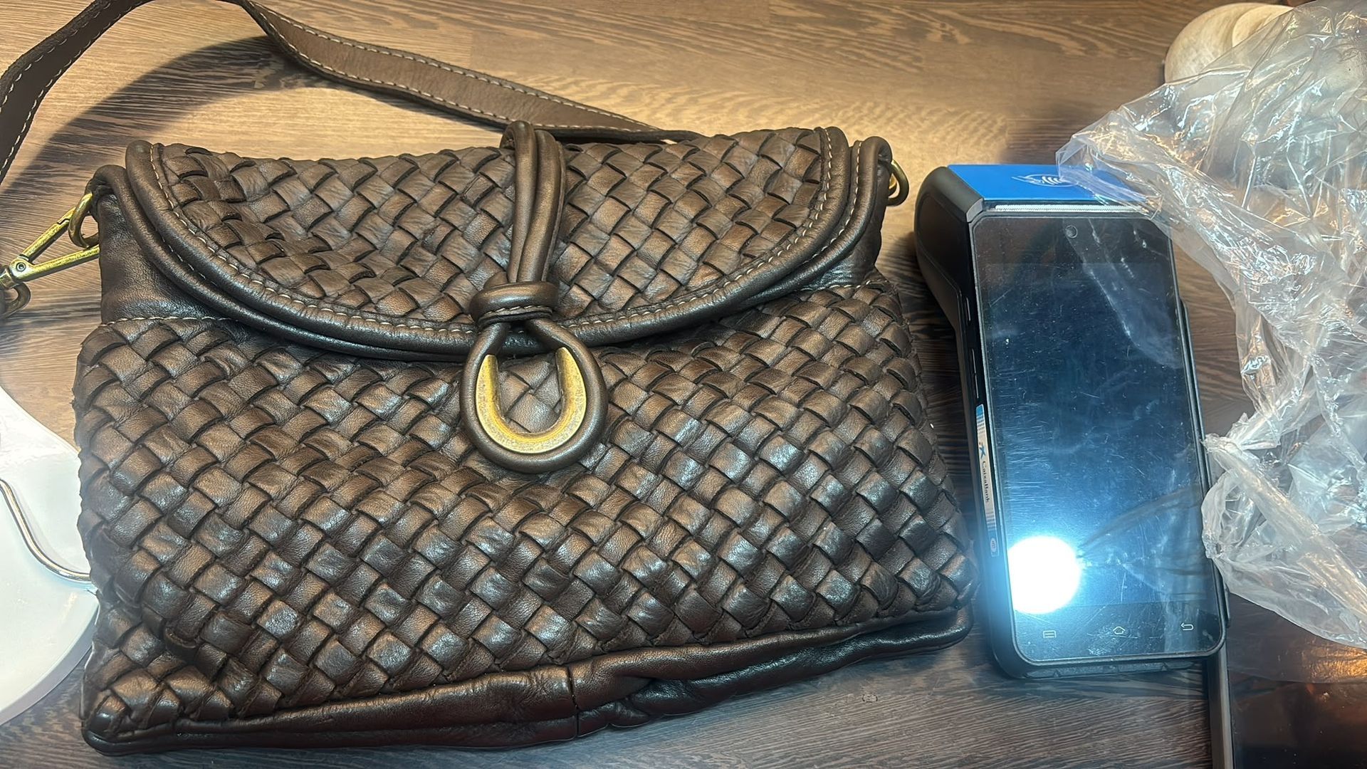 Bolso de mano negro tejido junto a un teléfono inteligente sobre una superficie de tela beige.