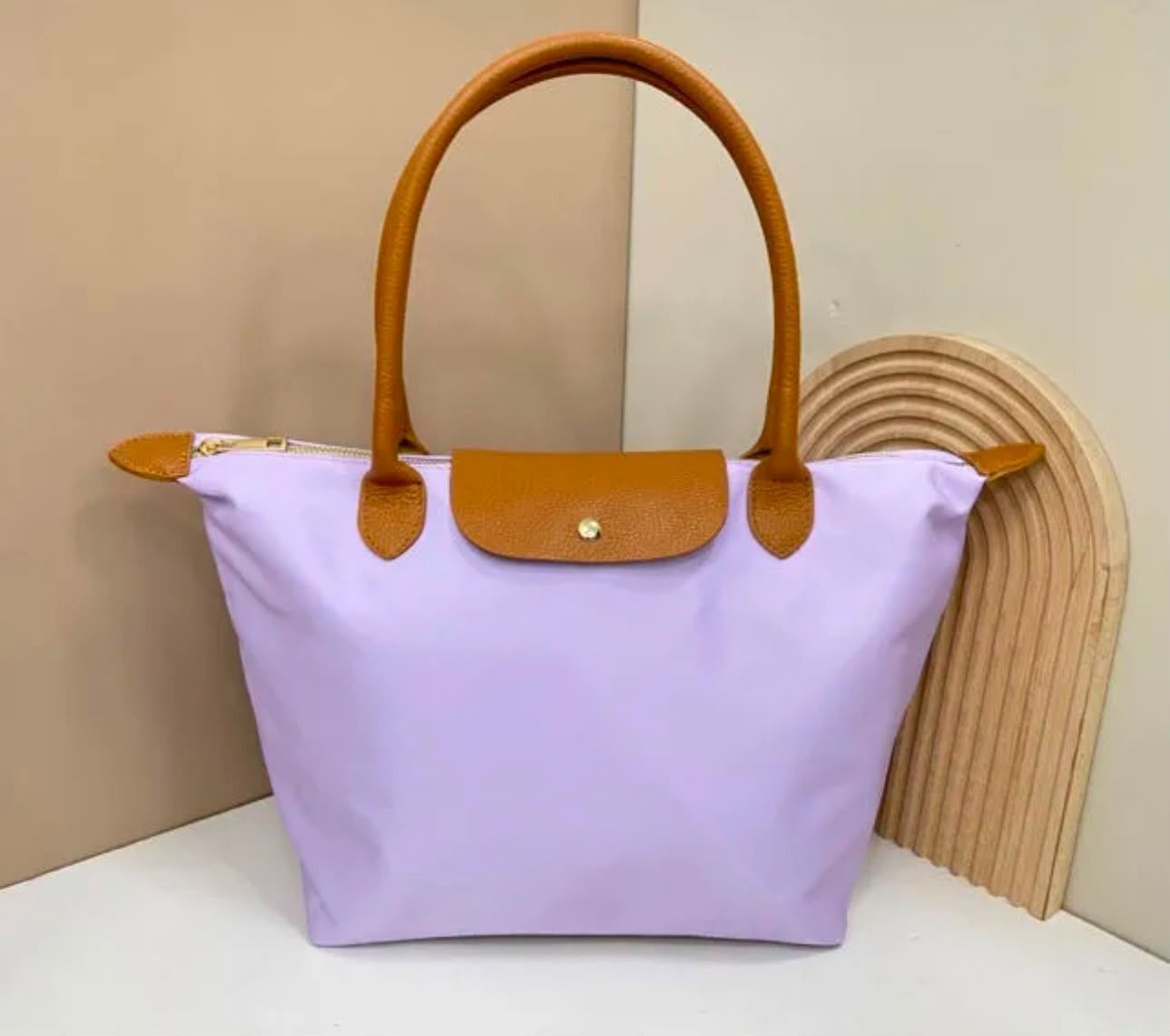 Bolso tote color lavanda con asas y solapa color tostado, exhibido sobre un fondo beige.