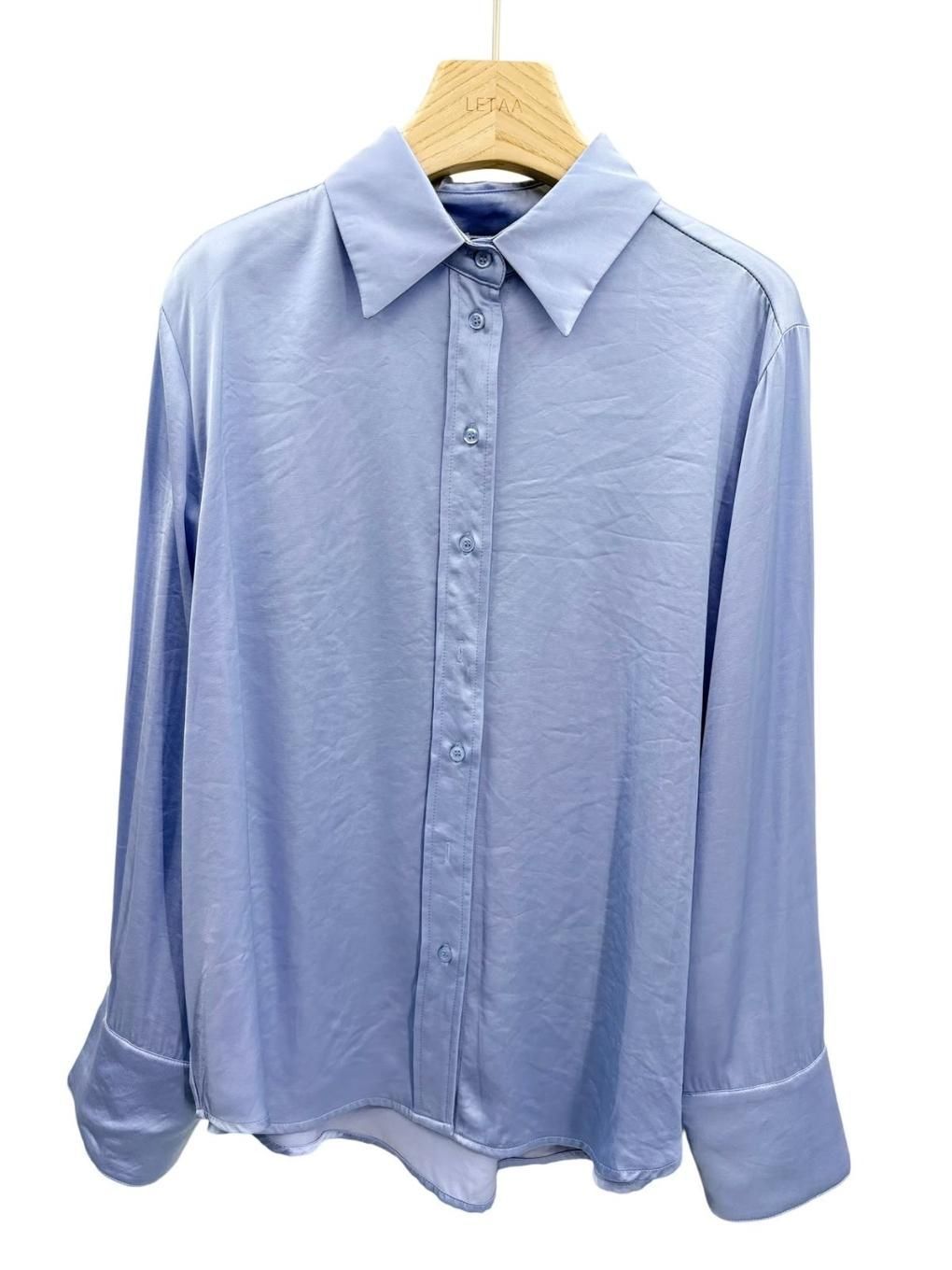 Camisa azul claro de manga larga con botones, colgada en una percha, vista frontal.