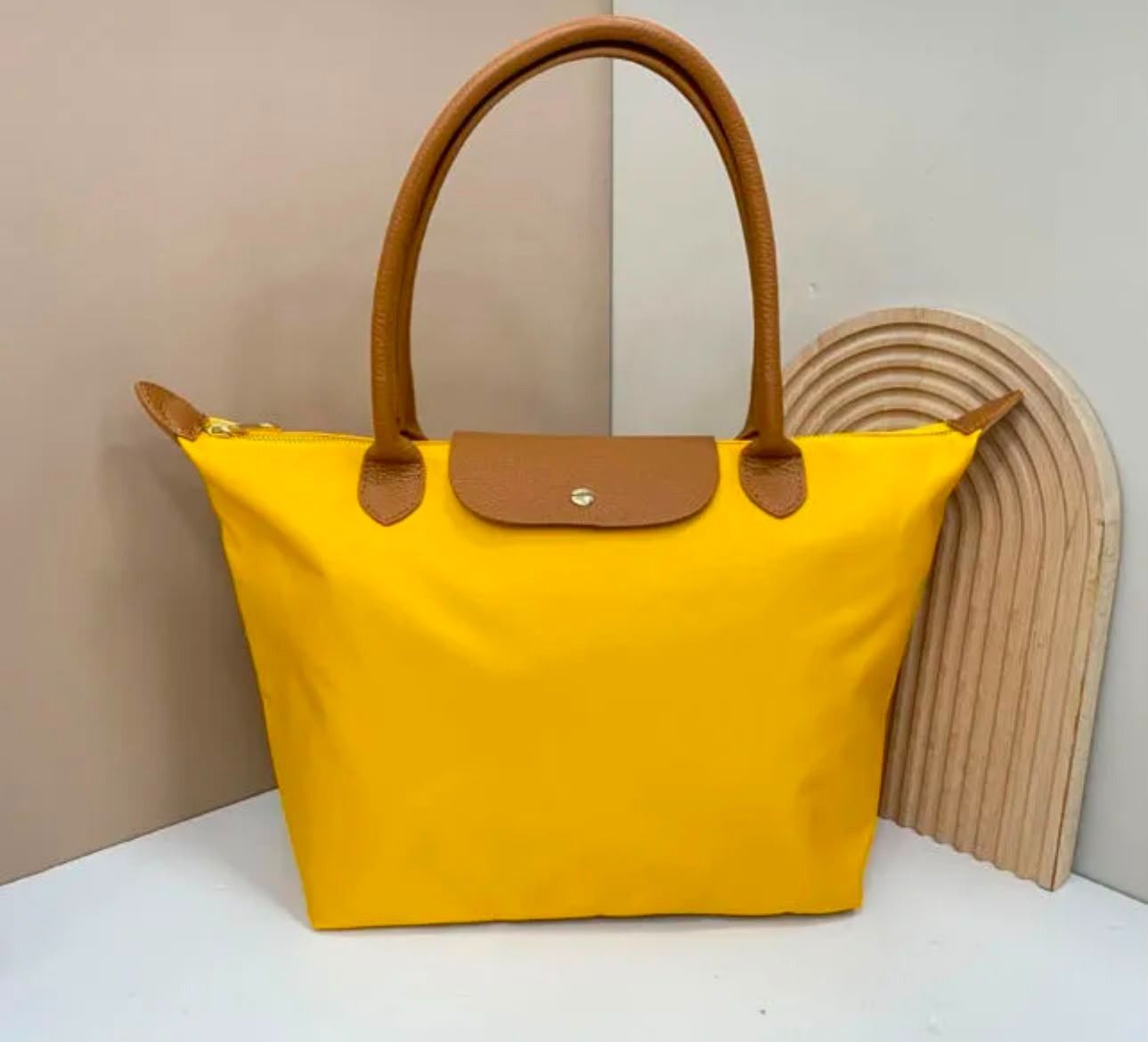 Bolso de mano amarillo con asas y ribetes marrones sobre una superficie blanca contra una pared beige.