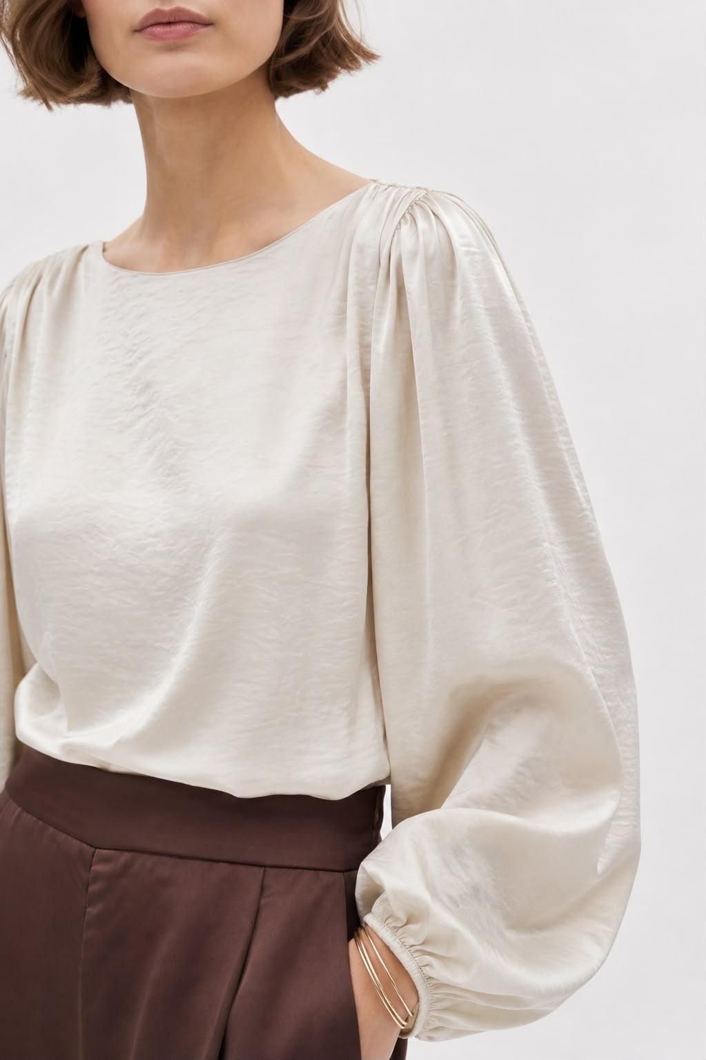 Mujer con blusa color crema de mangas abullonadas y falda marrón sobre un fondo claro.