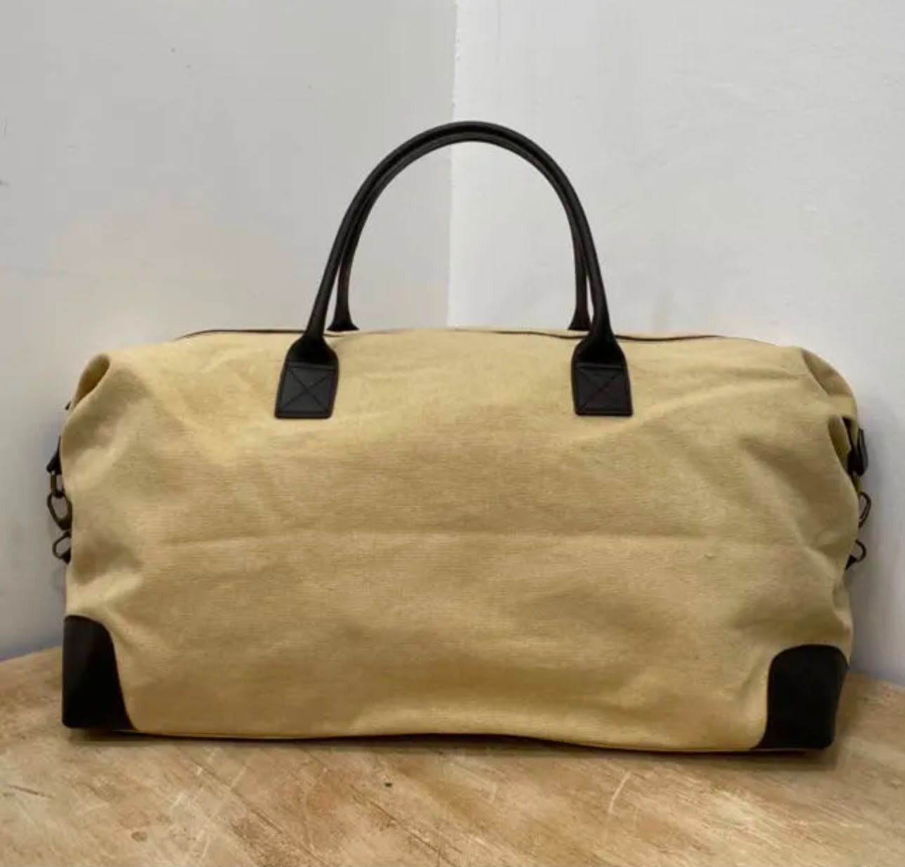 Bolsa de lona color beige con asas negras y ribete en las esquinas sobre una superficie de madera.