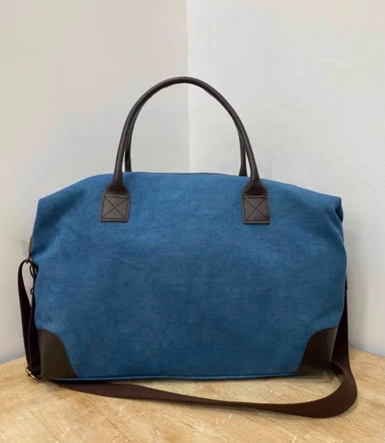 Bolsa de lona azul con asas y correa para el hombro marrones sobre un suelo claro.
