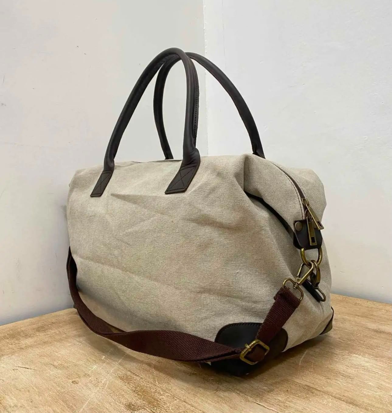 Bolsa de lona beige con asas negras y correa marrón sobre una mesa de madera contra una pared blanca.