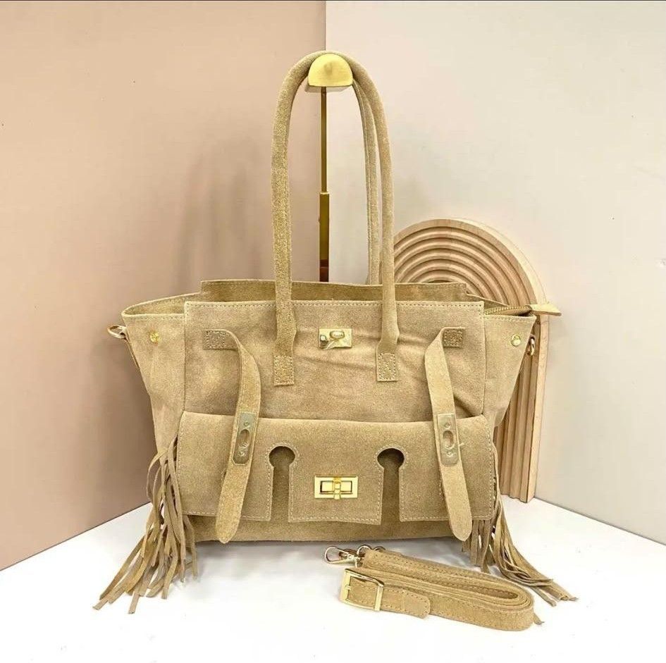Bolso de mano de ante color camel con flecos, correa larga y monedero redondo tejido sobre fondo beige.