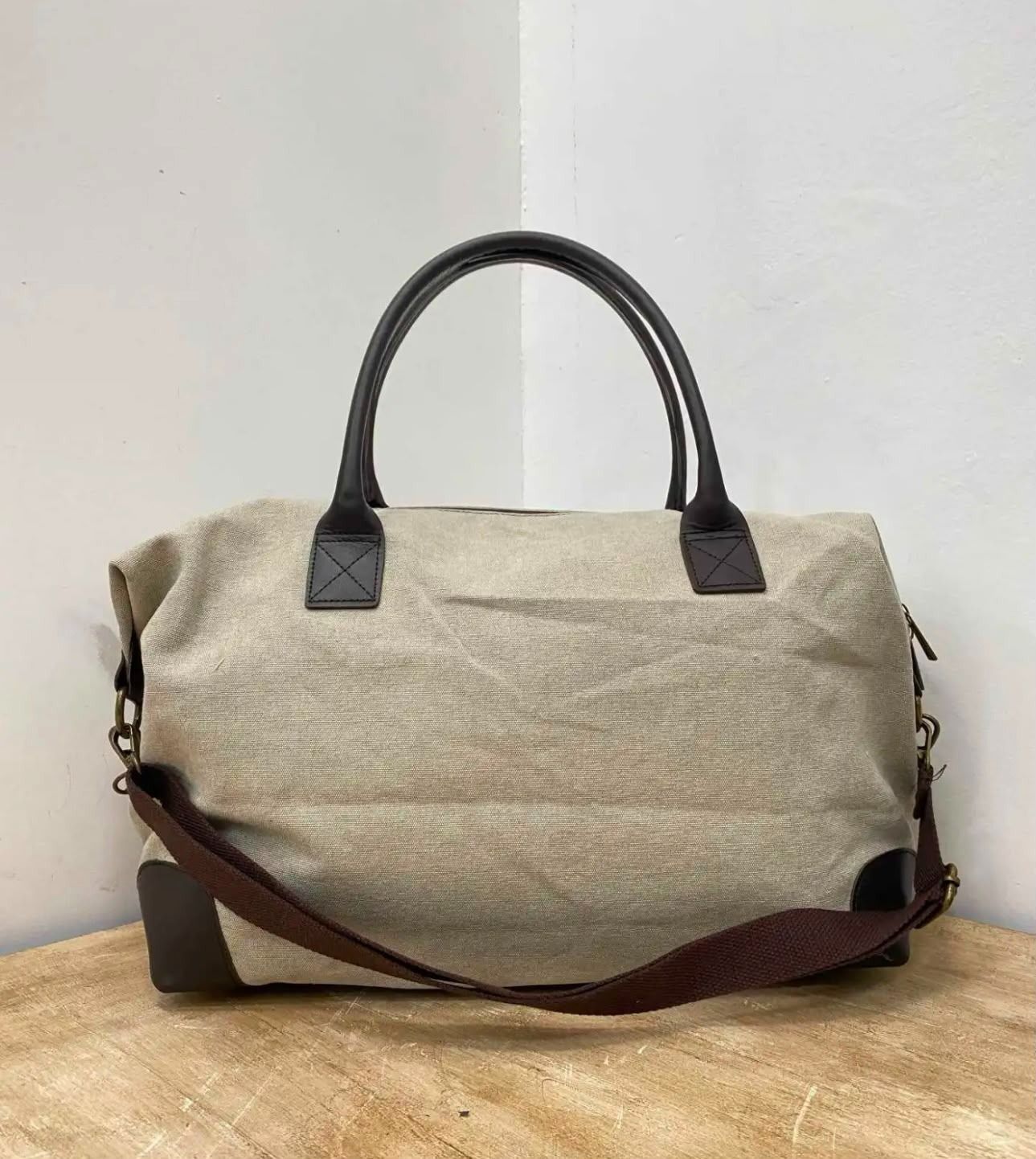 Bolsa de lona beige con asas negras y correa de hombro marrón sobre una mesa de madera.