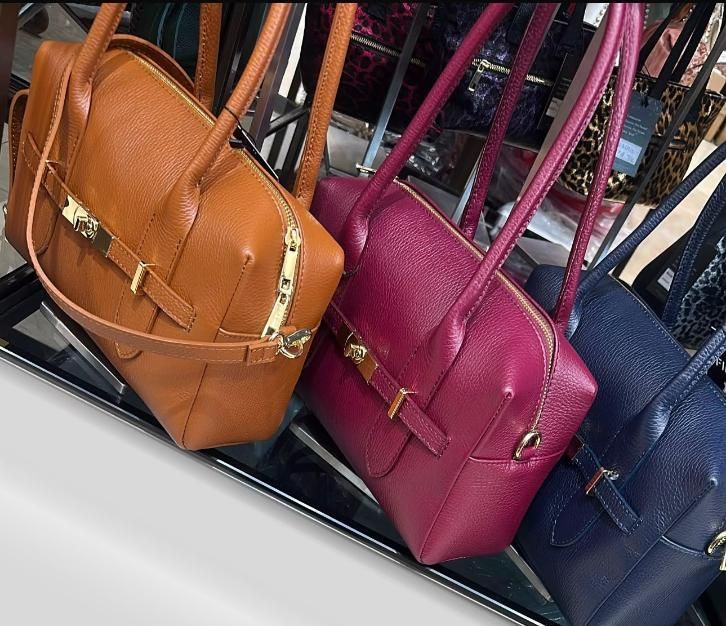 Tres bolsos de piel en color tostado, magenta y azul marino expuestos en un estante.