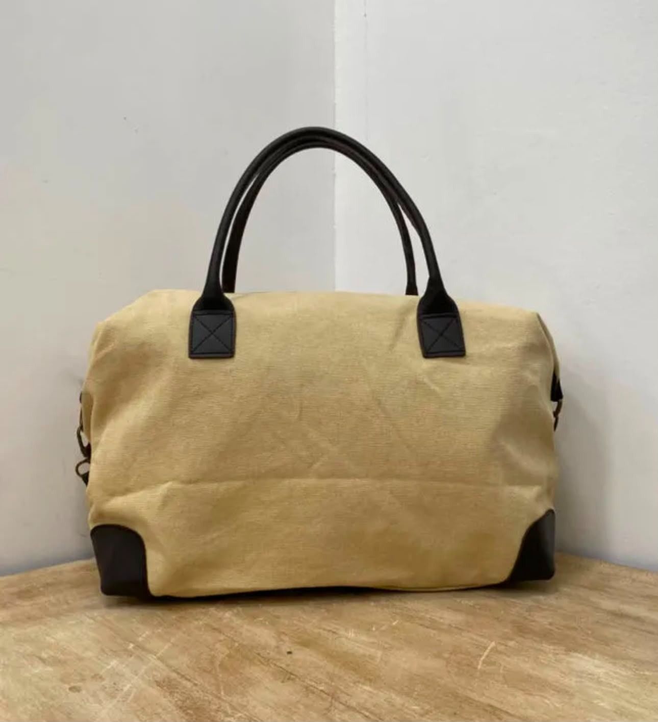 Bolsa de lona color canela con asas negras sobre una mesa de madera contra una pared lisa.