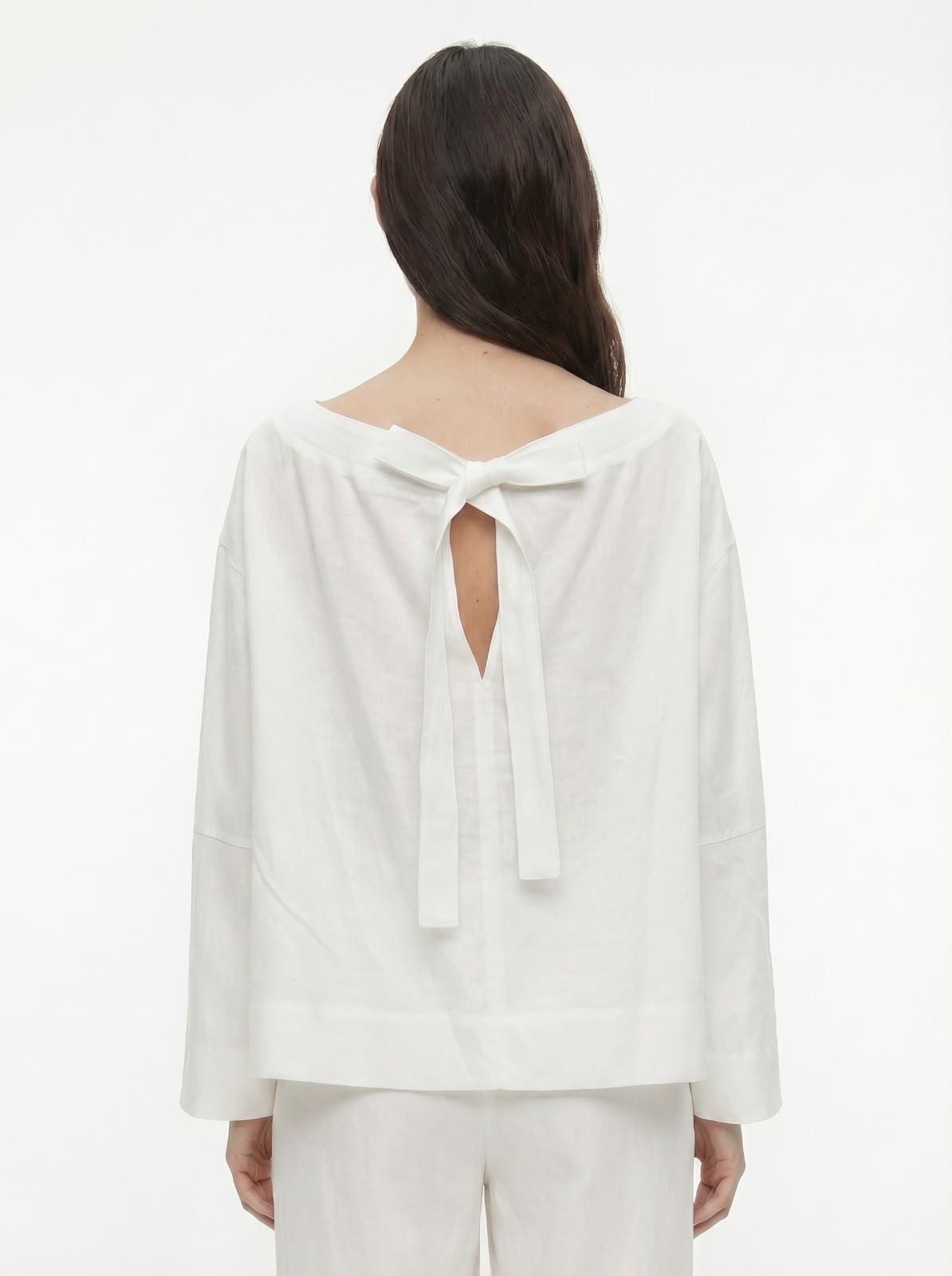 Persona con blusa blanca con espalda descubierta y detalle de lazo, vista desde atrás sobre un fondo liso.