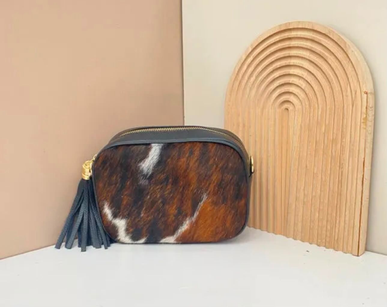 Bolsa de piel de vaca marrón y negra con borla negra junto a un arco decorativo de madera clara en un estante.