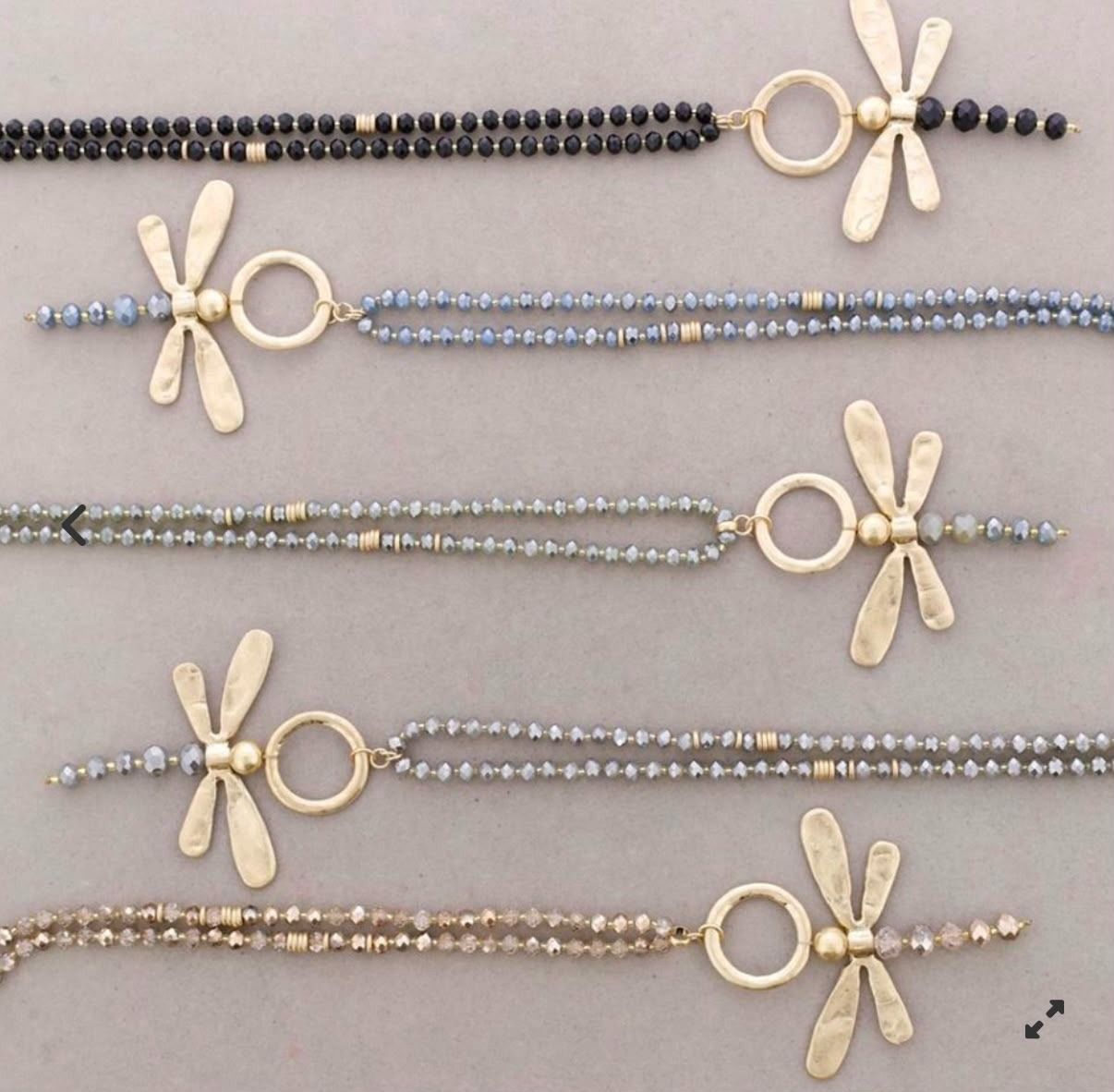 Cinco pulseras de cuentas en negro, azul, gris, color topo y beige con detalles de conchas y anillos dorados.