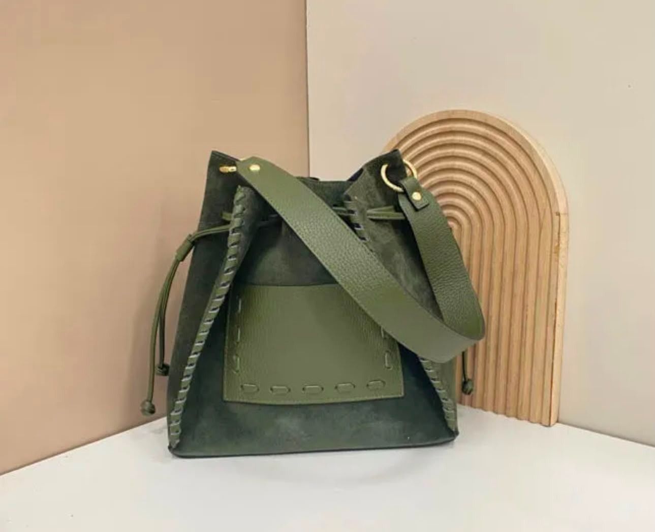 Bolso verde oliva con correa para el hombro sobre un estante junto a una pieza decorativa arqueada de color tostado.