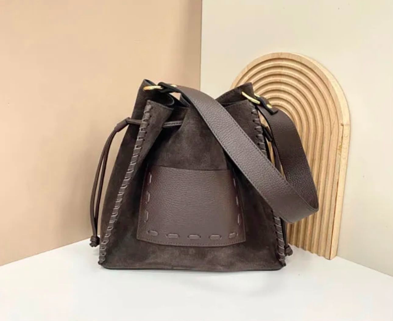 Bolso tipo cubo de cuero marrón oscuro con correas negras sobre una superficie blanca
