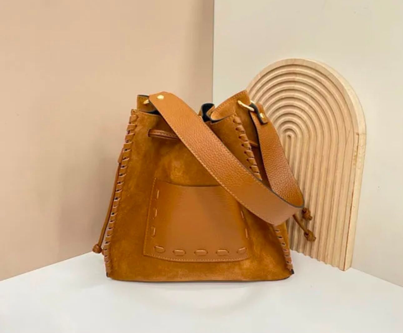 Bolso de mano de piel color camel con correa, situado junto a una pieza decorativa de madera acanalada