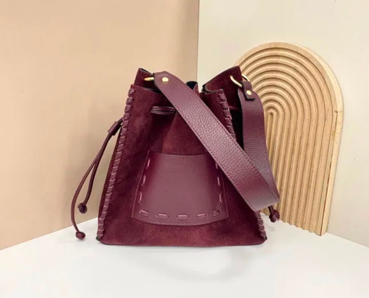 Bolso cubo de ante burdeos con correa larga, sobre una superficie blanca junto a un arco decorativo beige.