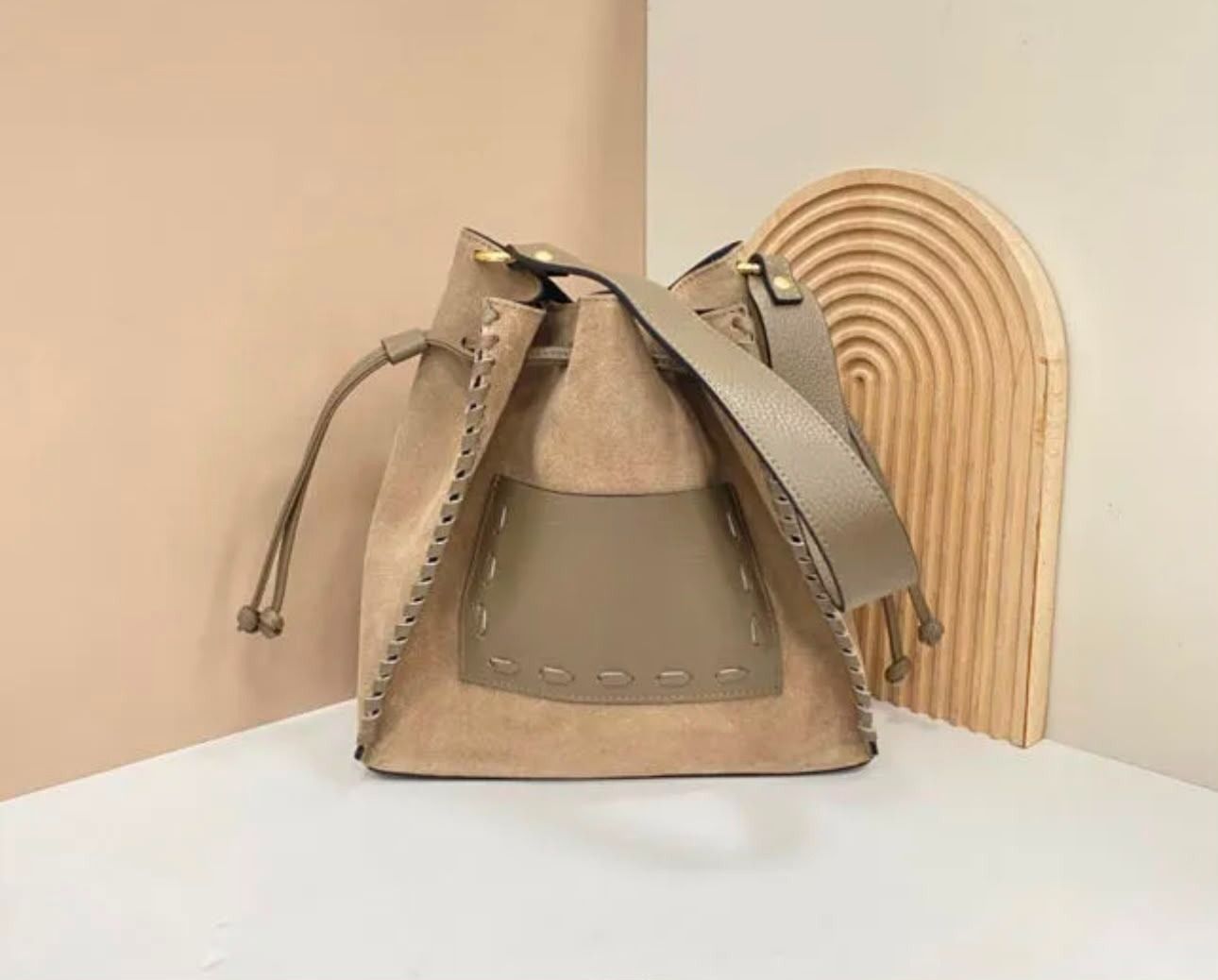 Un bolso de mano beige y gris con cordón ajustable sobre una mesa blanca junto a un elemento decorativo de arco de madera.