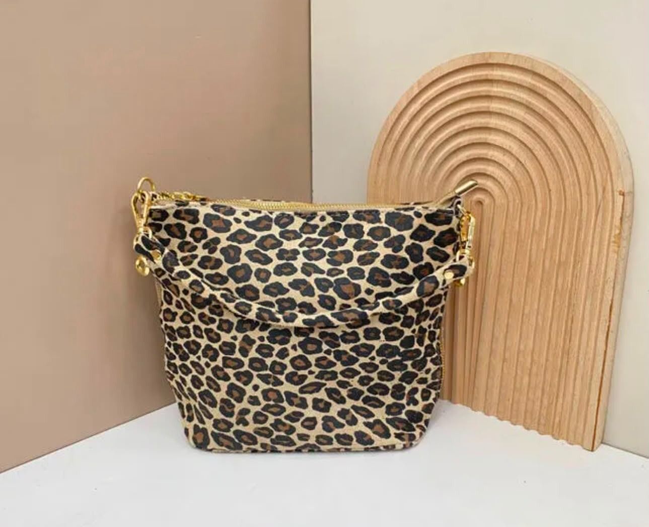 Un bolso tipo cubo con estampado de leopardo junto a un arco decorativo de madera acanalada sobre una superficie blanca.