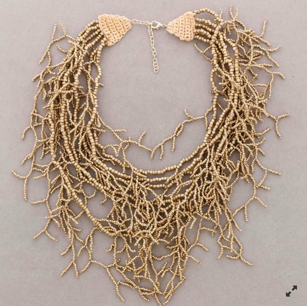 Collar de cuentas doradas con hilos superpuestos y detalles de cadena con flecos sobre un fondo gris.
