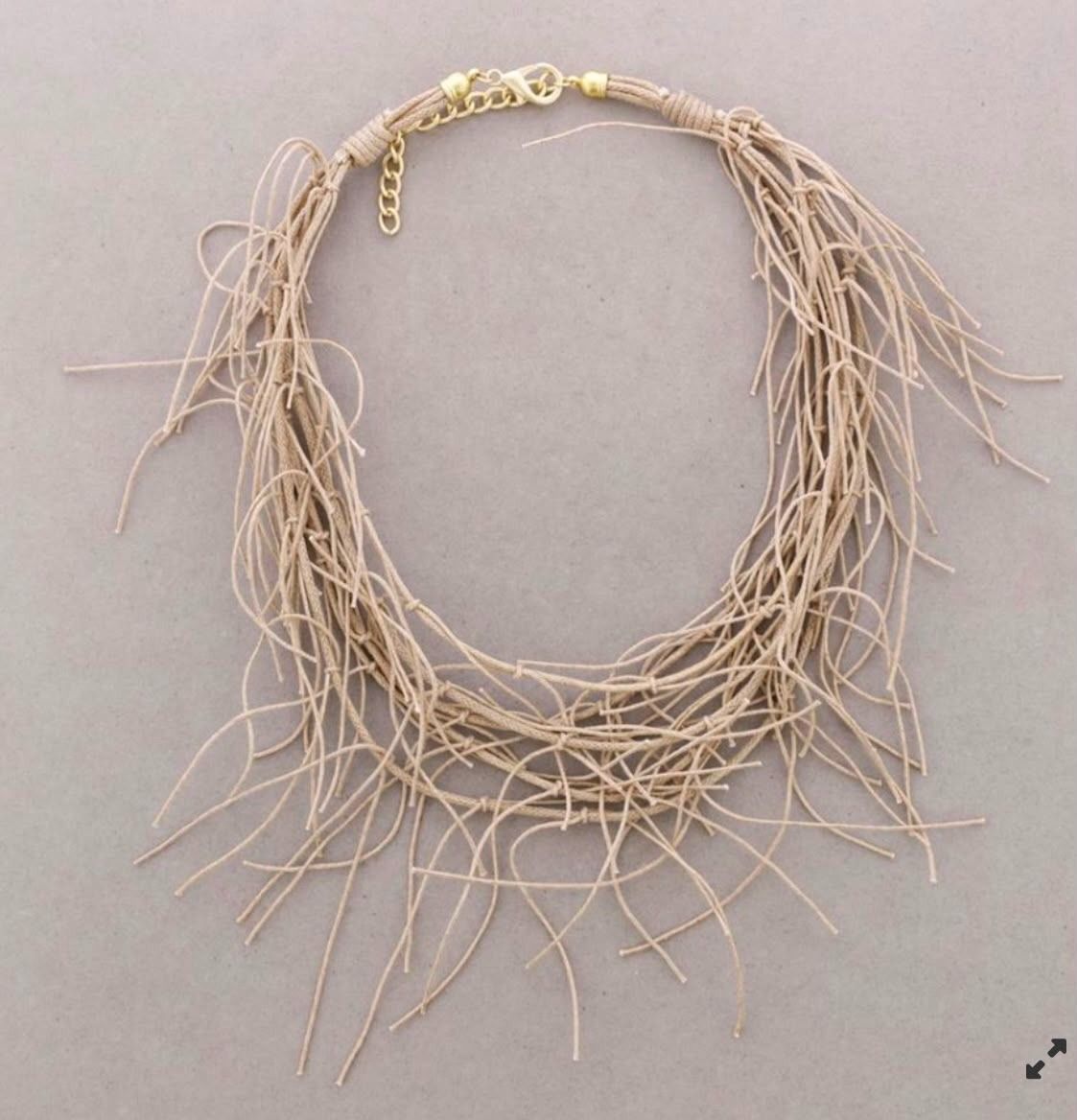 Collar tejido de color beige con hebras colgantes, extendido sobre un fondo gris.
