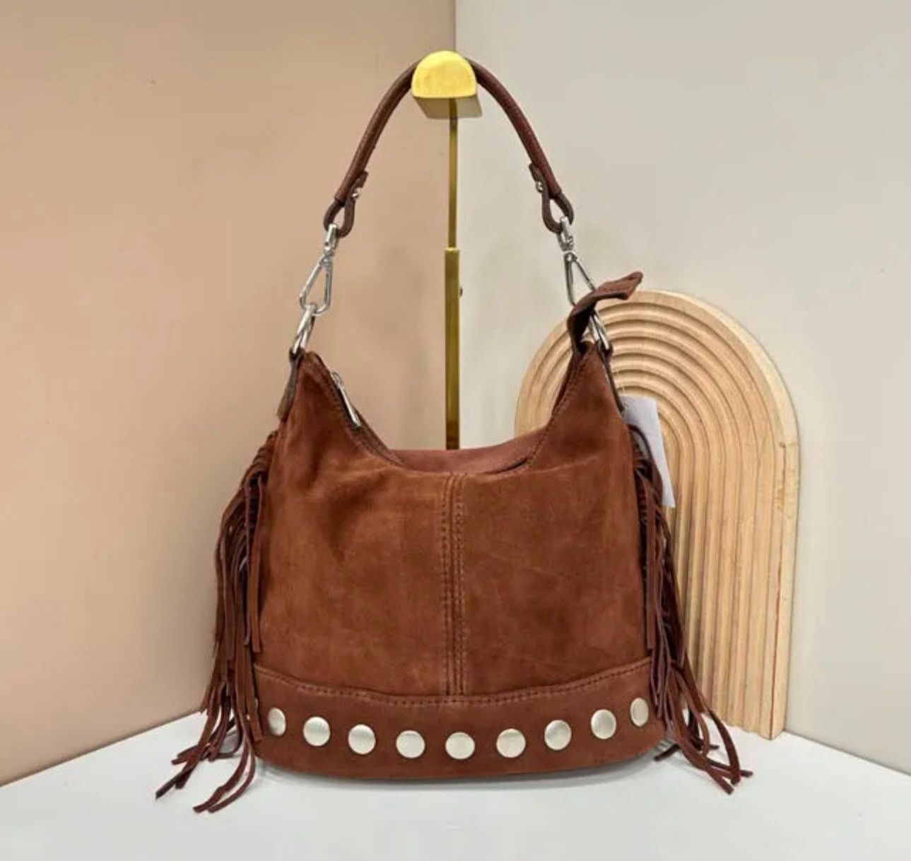 Bolso de mano de ante marrón con flecos y tachuelas plateadas, colocado junto a un arco de mimbre