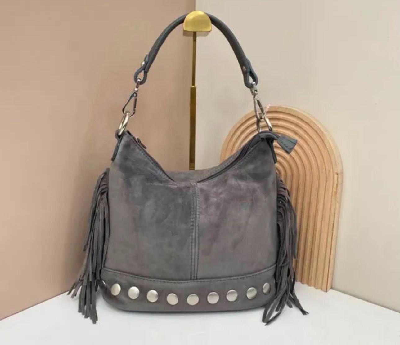 Bolso de mano de ante gris con flecos y tachuelas plateadas sobre un soporte, contra una pared beige.