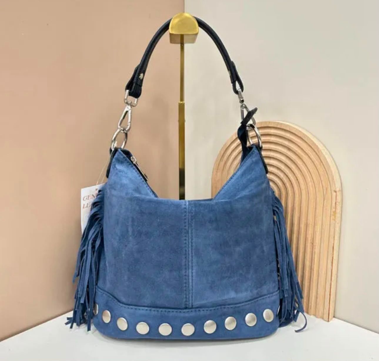Bolso de hombro de mezclilla azul con correas negras y tachuelas plateadas, expuesto en un soporte.