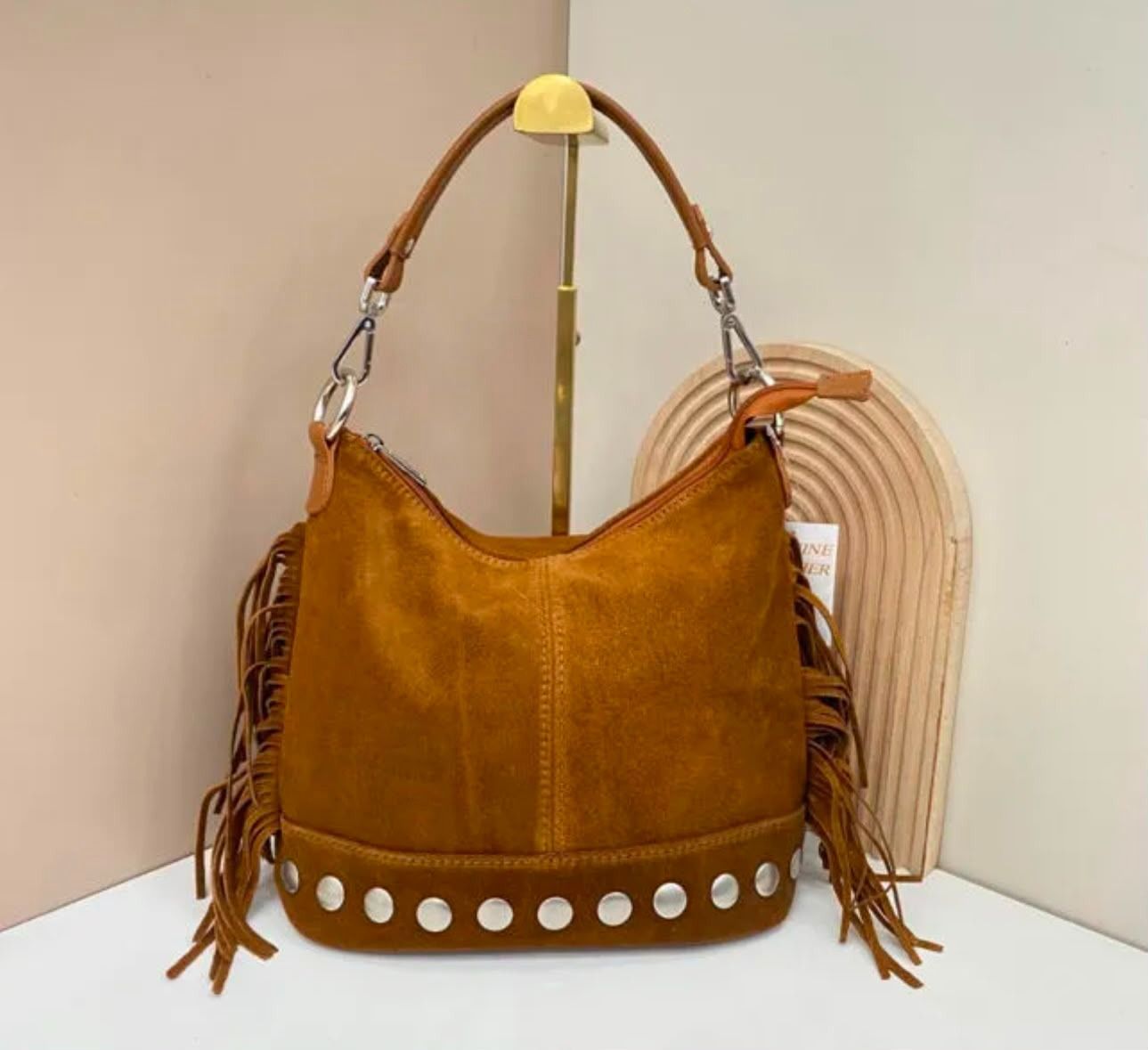 Bolso de mano de ante color camel con flecos y tachuelas plateadas, colgado en un expositor.