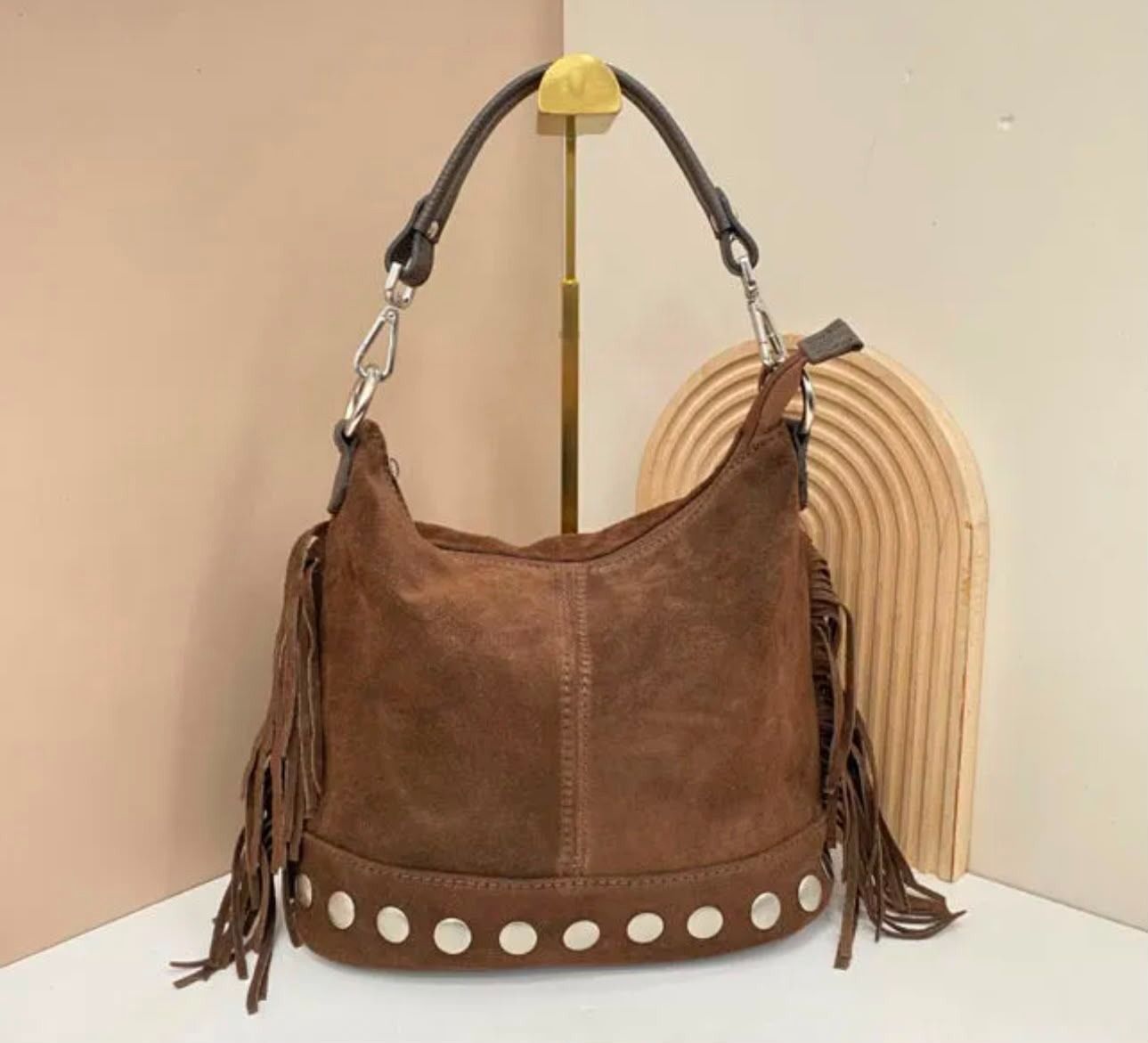 Bolso de mano marrón de ante con flecos, asas de anillas plateadas y tachuelas blancas, expuesto sobre un soporte.