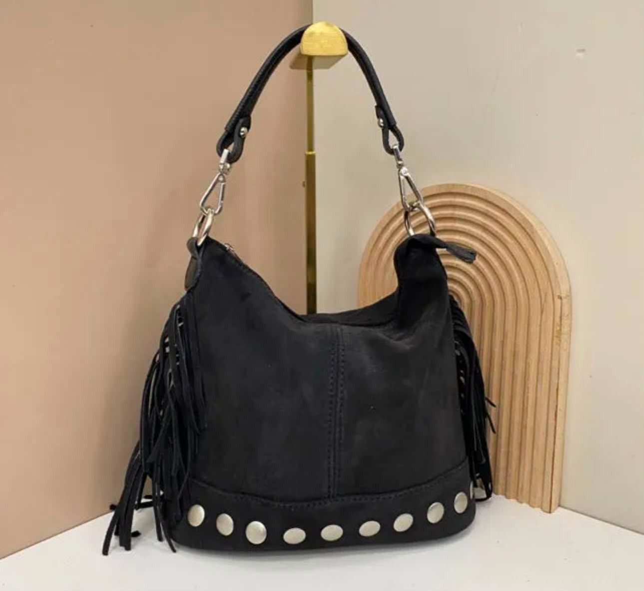 Bolso hobo negro con flecos, base con tachuelas plateadas y correa de cadena, expuesto sobre una mesa.