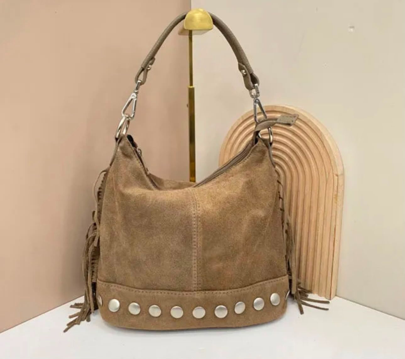 Bolso de hombro de ante color camel con tachuelas plateadas, mostrado sobre una superficie blanca contra una pared beige.