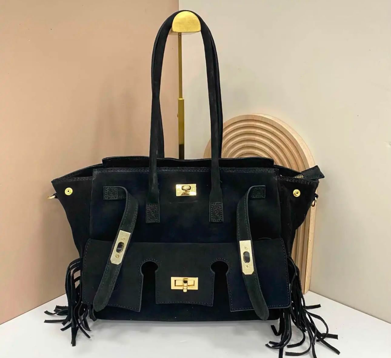 Bolso de mano de ante negro con herrajes dorados y borlas sobre una superficie blanca junto a una silla de mimbre.