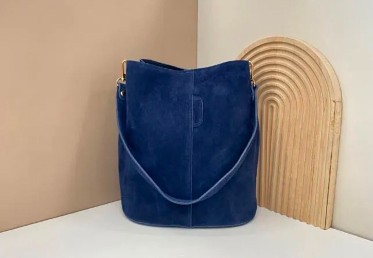 Un bolso de terciopelo azul sobre una superficie blanca junto a un panel decorativo tejido de color beige.
