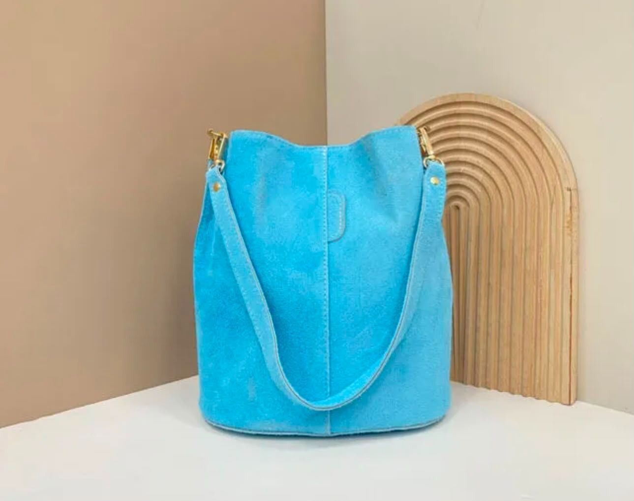 Un bolso de mano azul brillante con cremalleras doradas sobre una mesa blanca, con una pared beige y una silla tejida.