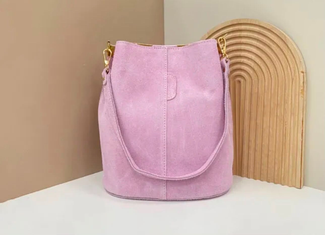 Un bolso cubo de ante rosa sobre una mesa blanca contra una pared beige con una pieza decorativa de madera curvada.
