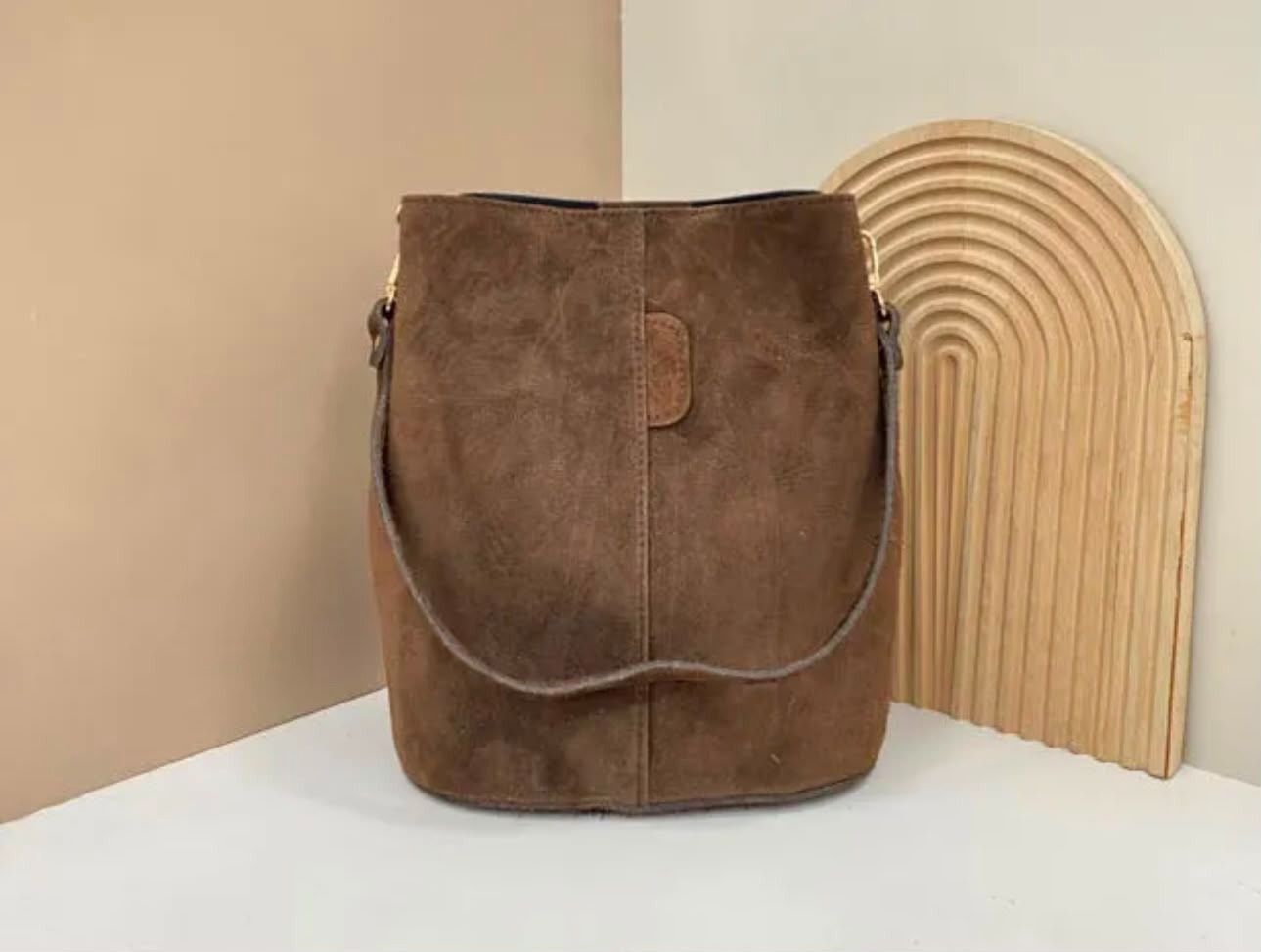 Bolso bandolera de ante marrón sobre una superficie blanca junto a una pieza decorativa de madera curvada.