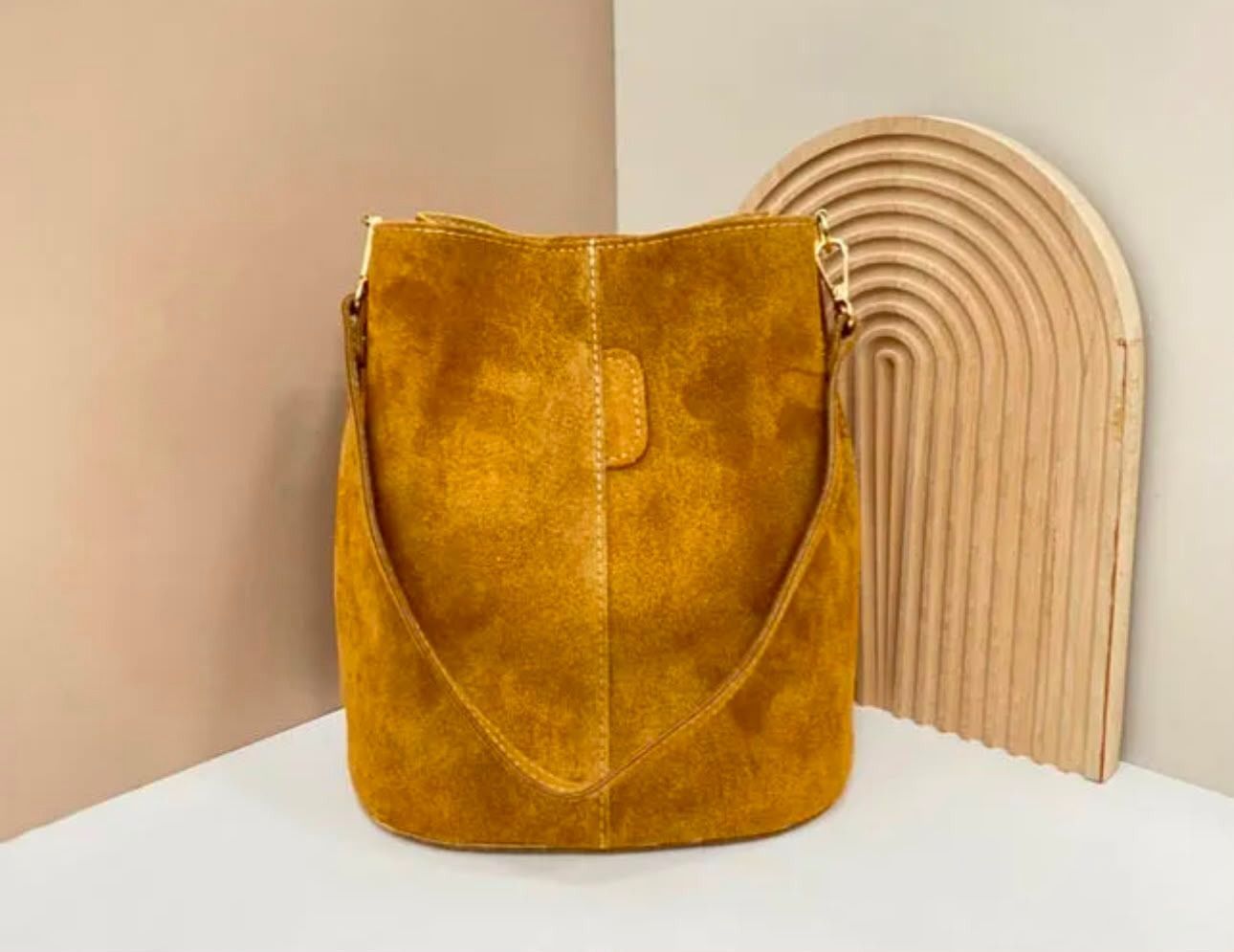 Un bolso de ante color mostaza sobre una superficie blanca, junto a una pieza decorativa de madera arqueada de color beige.