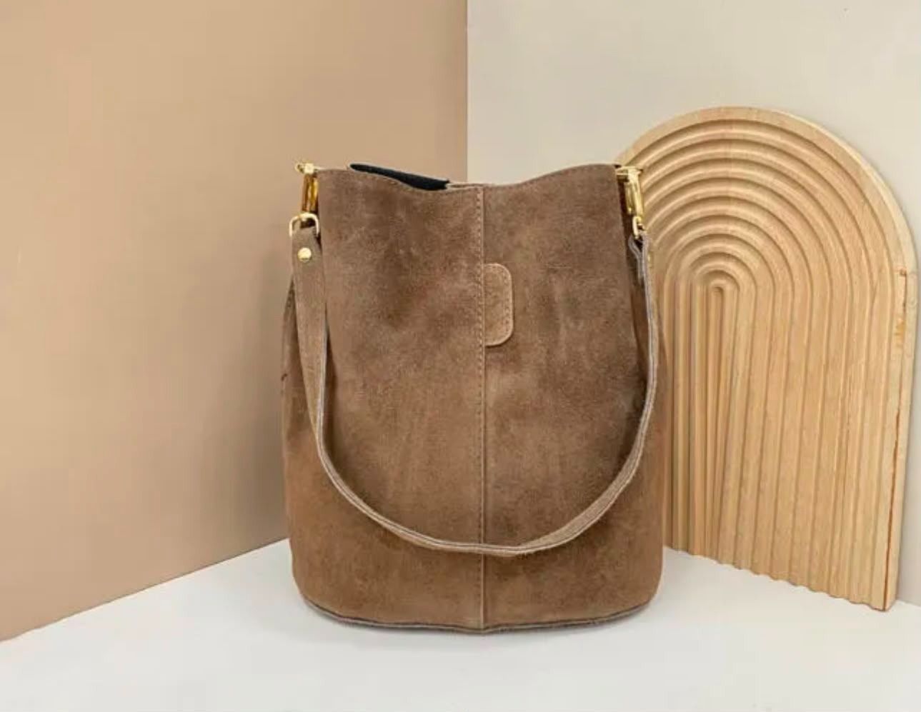 Bolso tipo cubo de ante marrón con correa curvada junto a una pieza decorativa de arco de madera sobre una superficie beige.