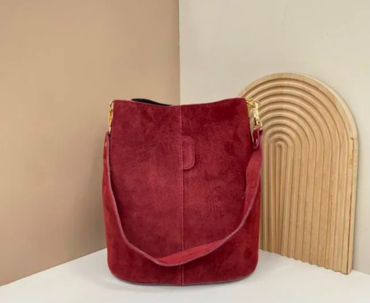 Un bolso tipo cubo de gamuza roja sobre una superficie blanca, junto a una pieza decorativa de madera curvada.