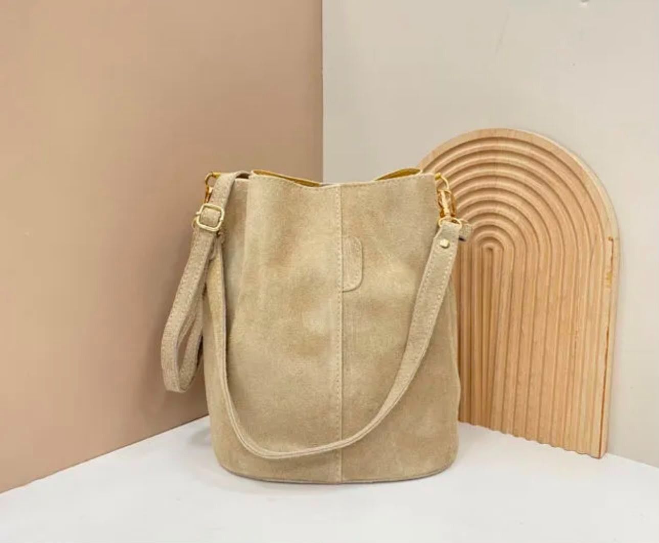 Bolso tipo cubo de ante beige con herrajes de correa dorados sobre una superficie blanca