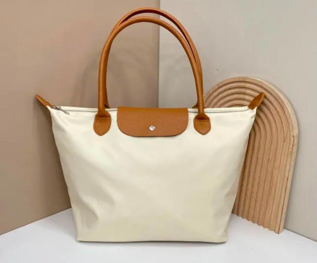 Bolso tote color crema con asas y ribetes de cuero marrón claro sobre un fondo beige.