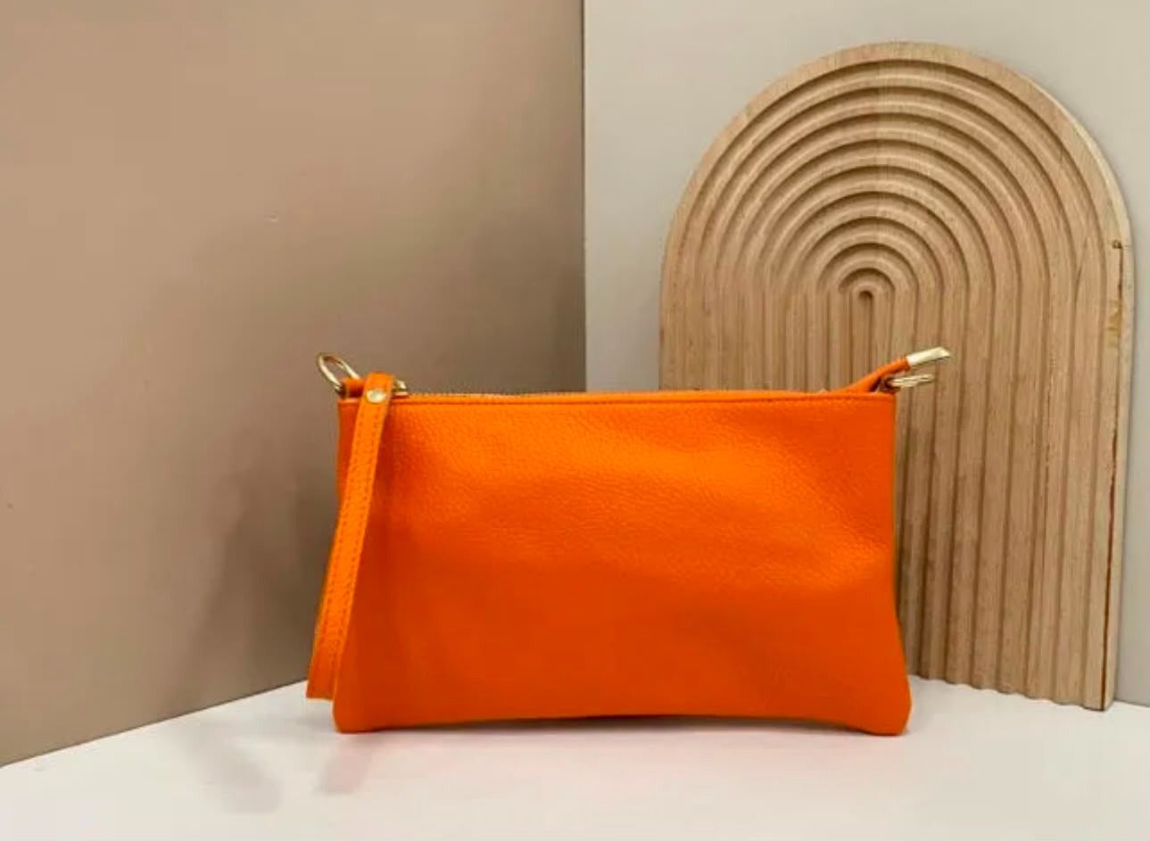 Un estuche naranja con cremallera sobre una superficie blanca, junto a una pieza decorativa arqueada de color beige.